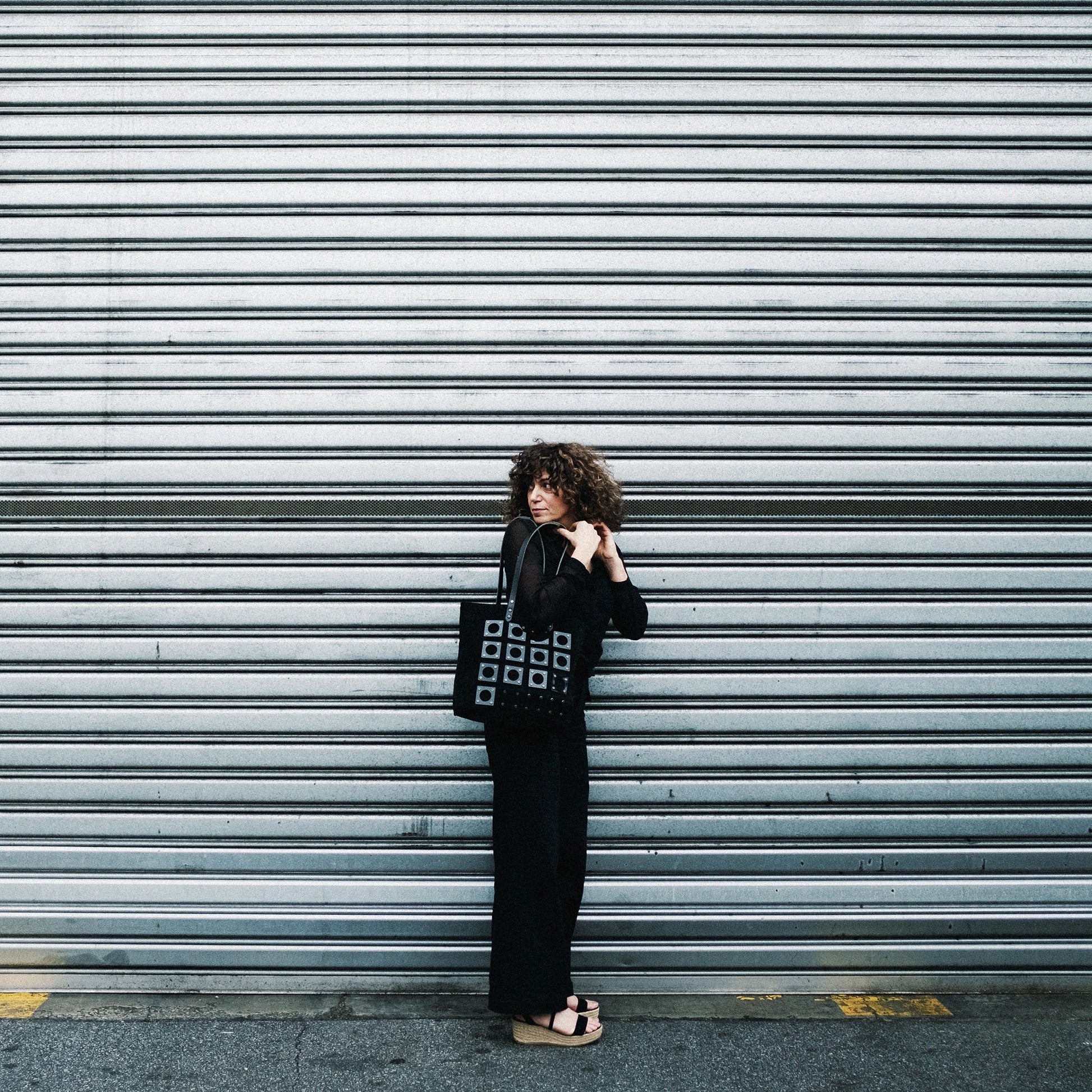 METANoIA black recycled leather medium handbag with square and hollowed circle black acrylic forms fashioned into a repetitive fashion. Model featured leaning on industrial metal wall, looking over her shoulder with bag over her shoulder.