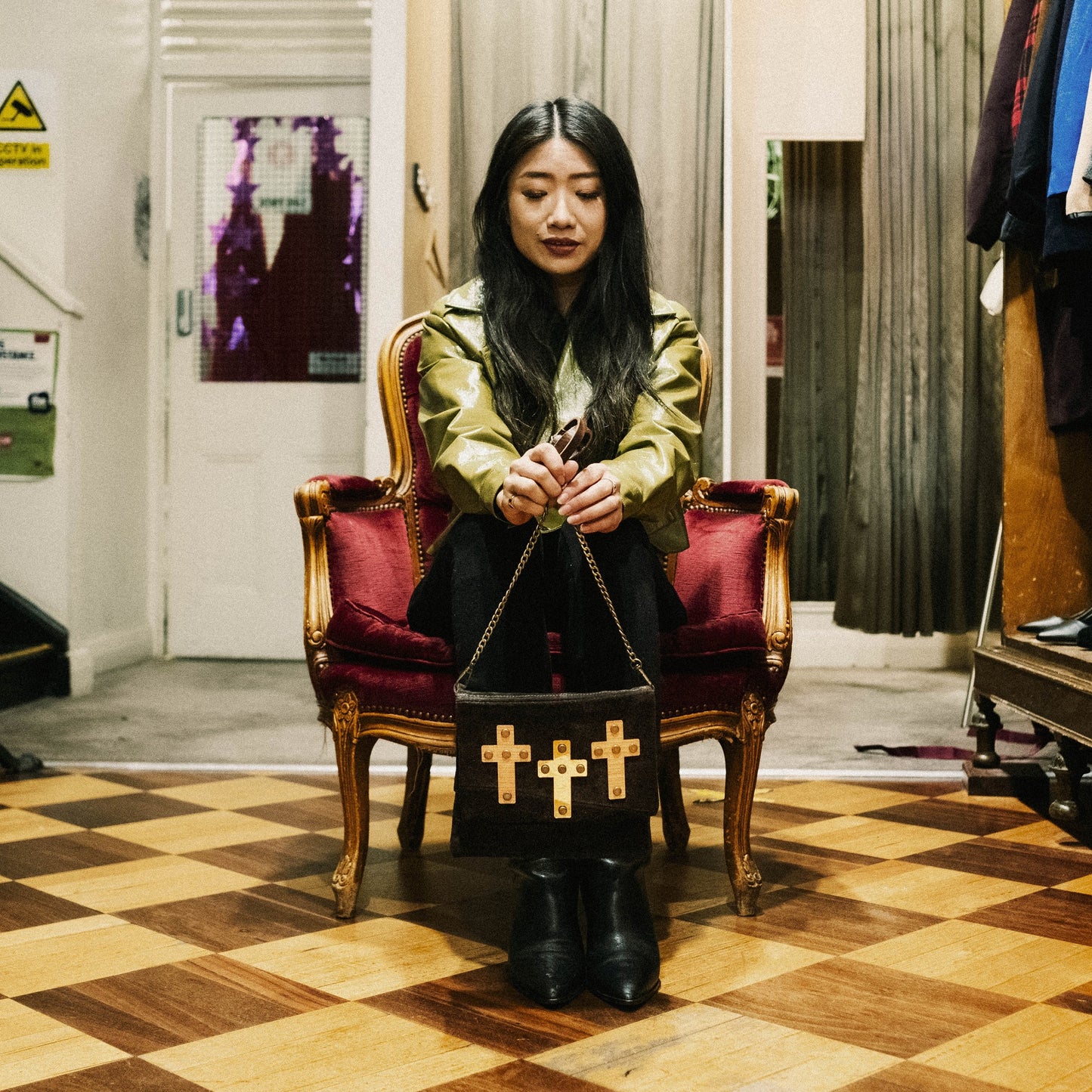 METANoIA The Cross Black recycled leather small handbag with cross shaped bamboo, walnut and metallic acrylic forms. Model featured wearing a high sheen green jacket, seated on a burgundy vintage velvet chair with the small cross bag infront of her.