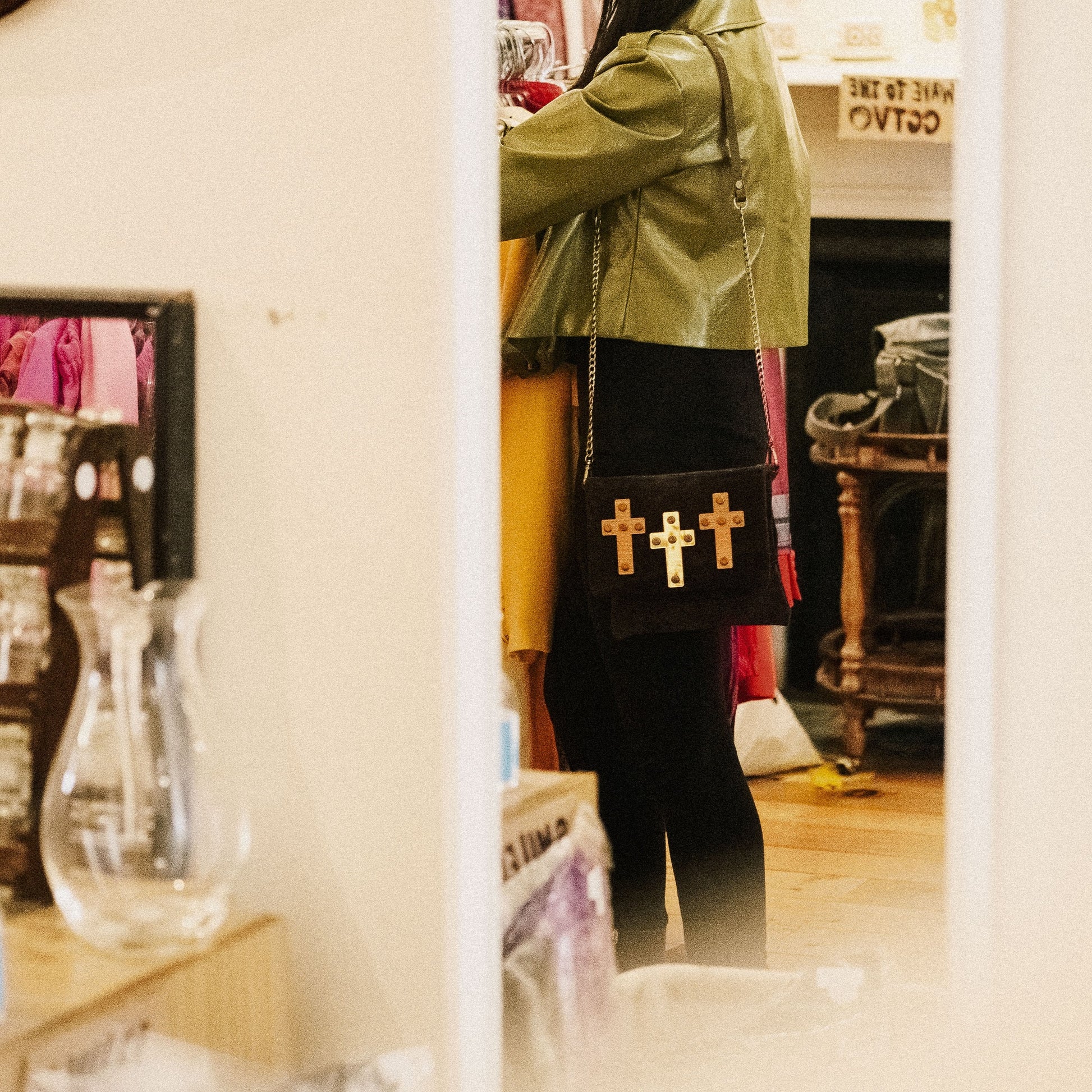 METANoIA The Cross Black recycled leather small handbag with cross shaped bamboo, walnut and metallic acrylic forms. Model featured wearing a high sheen green jacket, browsing through vintage clothing with the small cross bag beside her.