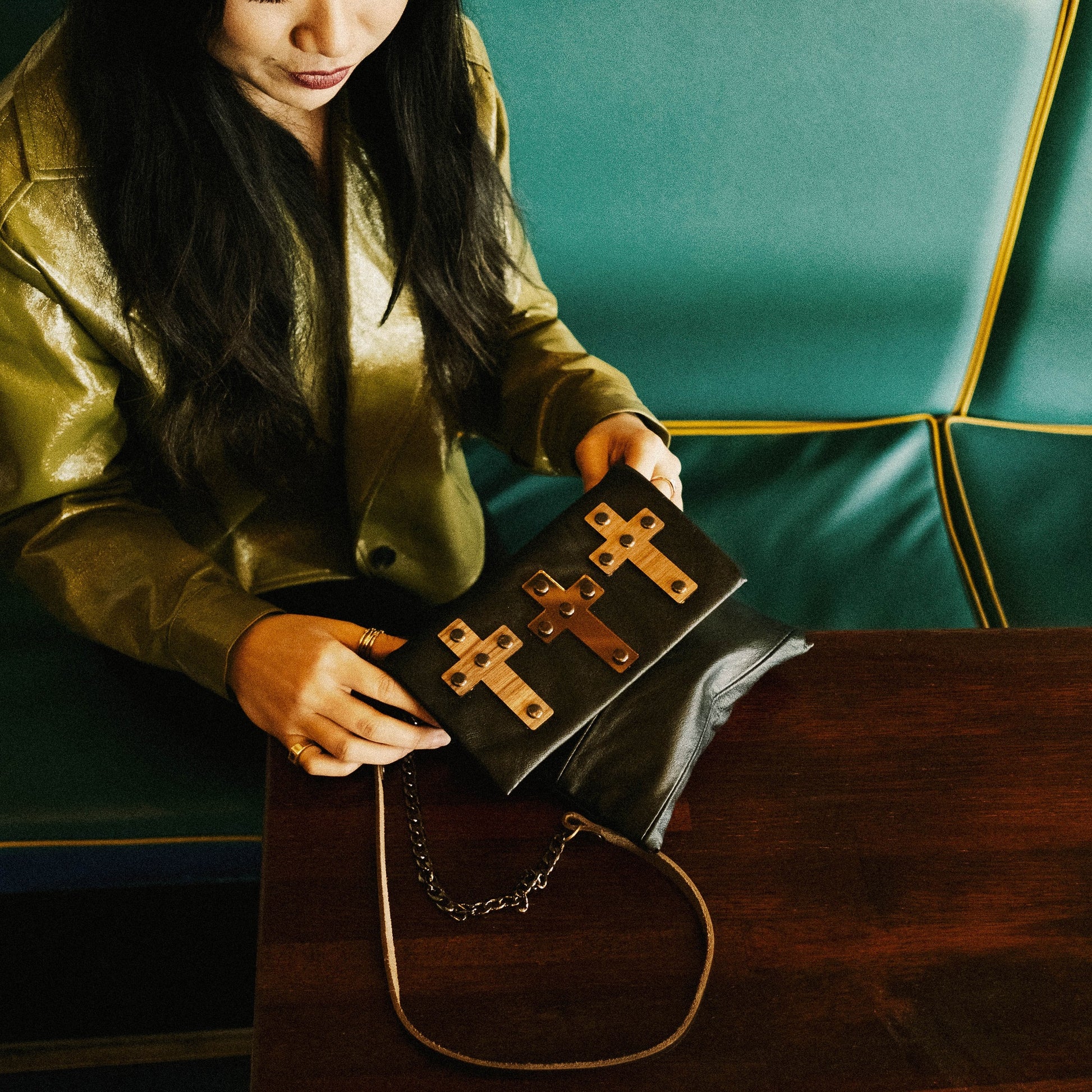 METANoIA The Cross Chocolate recycled leather small handbag with cross shaped bamboo, walnut and metallic acrylic forms. Model featured with cross bag placed on rich cherry wooden table, contrasted with green vinyl seating.