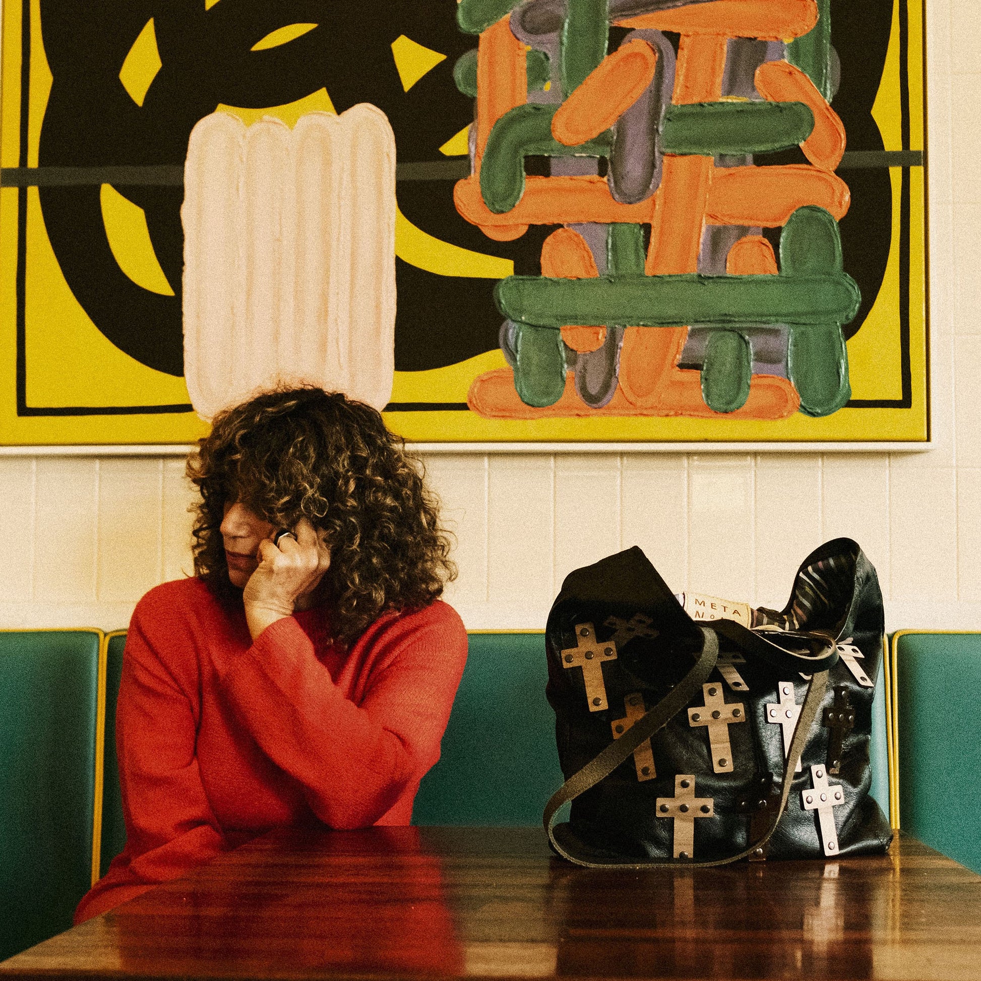 METANoIA The Cross Black recycled leather tote handbag with cross shaped bamboo, walnut and metallic acrylic forms. Model seated infront of abstract artwork with bag on the table by her side.