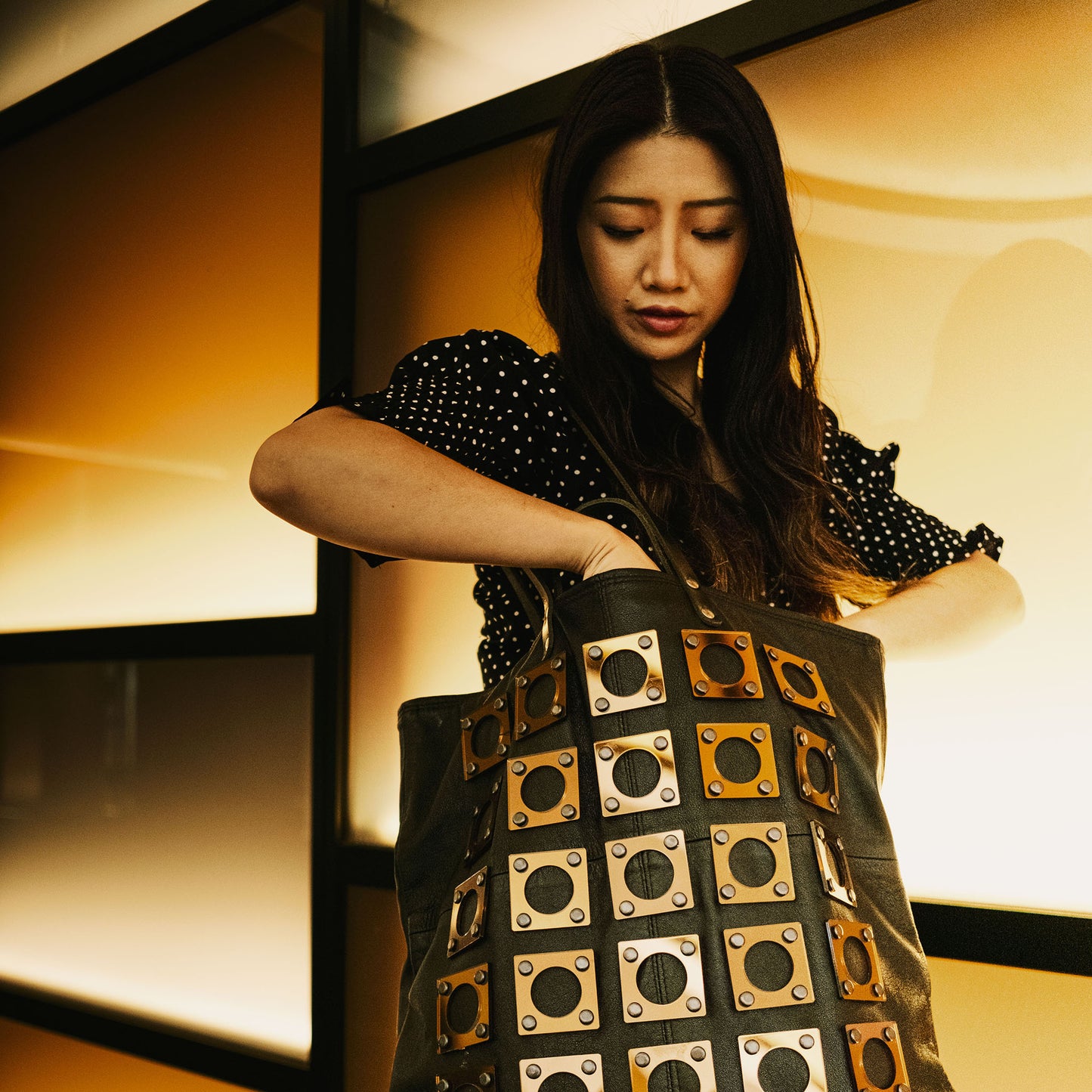 METANoIA black recycled leather tote handbag with square and hollowed circle copper acrylic forms fashioned into a repeative fashion. Model standing in train station reaching into the tote bag.