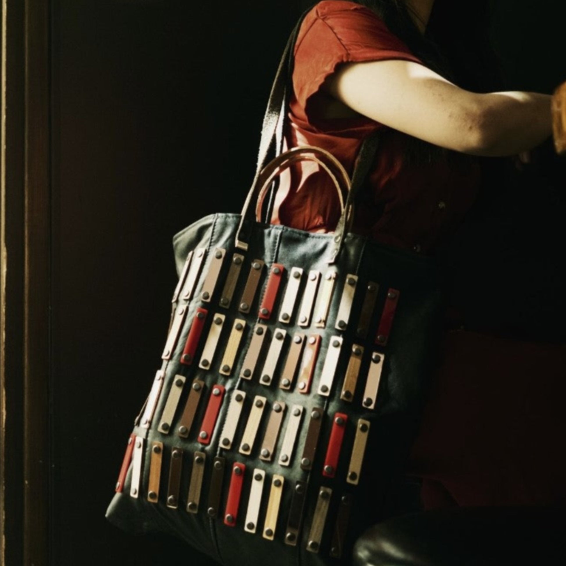 METANoIA black recycled leather tote handbag with acrylic and wooden vertical accents studded in a repeatitive fashion in an array of natural wooden brown colours, hints of red and copper. This large tote bag is featured on a model seated on a bench.