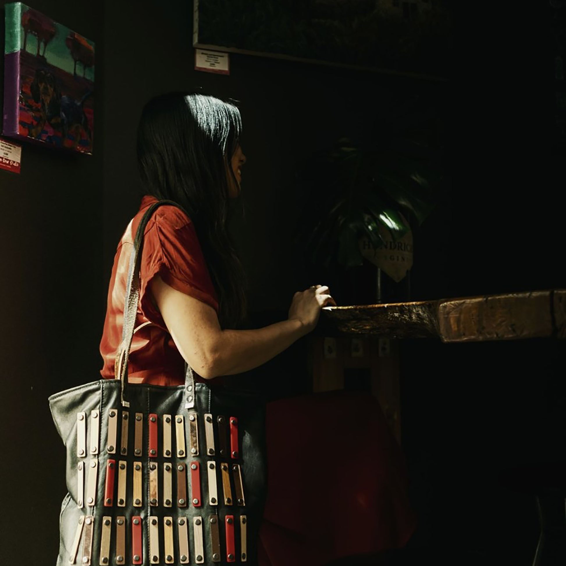 METANoIA black recycled leather tote handbag with acrylic and wooden vertical accents studded in a repeatitive fashion in an array of natural wooden brown colours, hints of red and copper. Featured on model seated and leaning on a wooden bench.