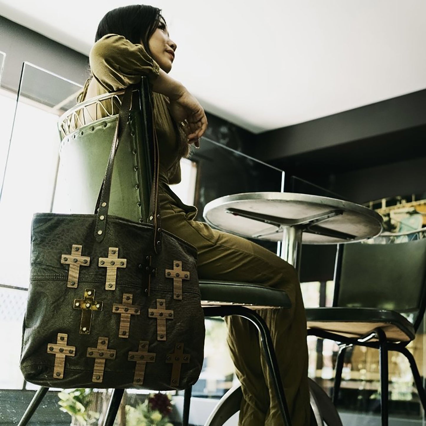 METANoIA The Cross Chocolate recycled leather medium handbag with cross shaped bamboo, walnut and metallic acrylic forms. Model seated on a vintage green leather chair with bag hanging on chair.