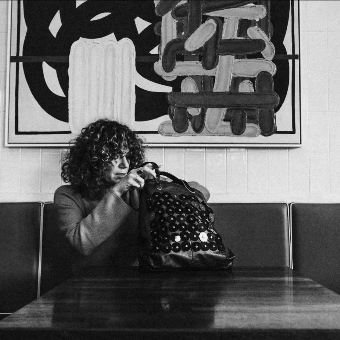METANoIA black recycled leather medium handbag with black circle acrylic forms fashioned into a repeative fashion with a smaller circle overlay on each form. Model featured seated at a restaurant with the bag placed on the table.