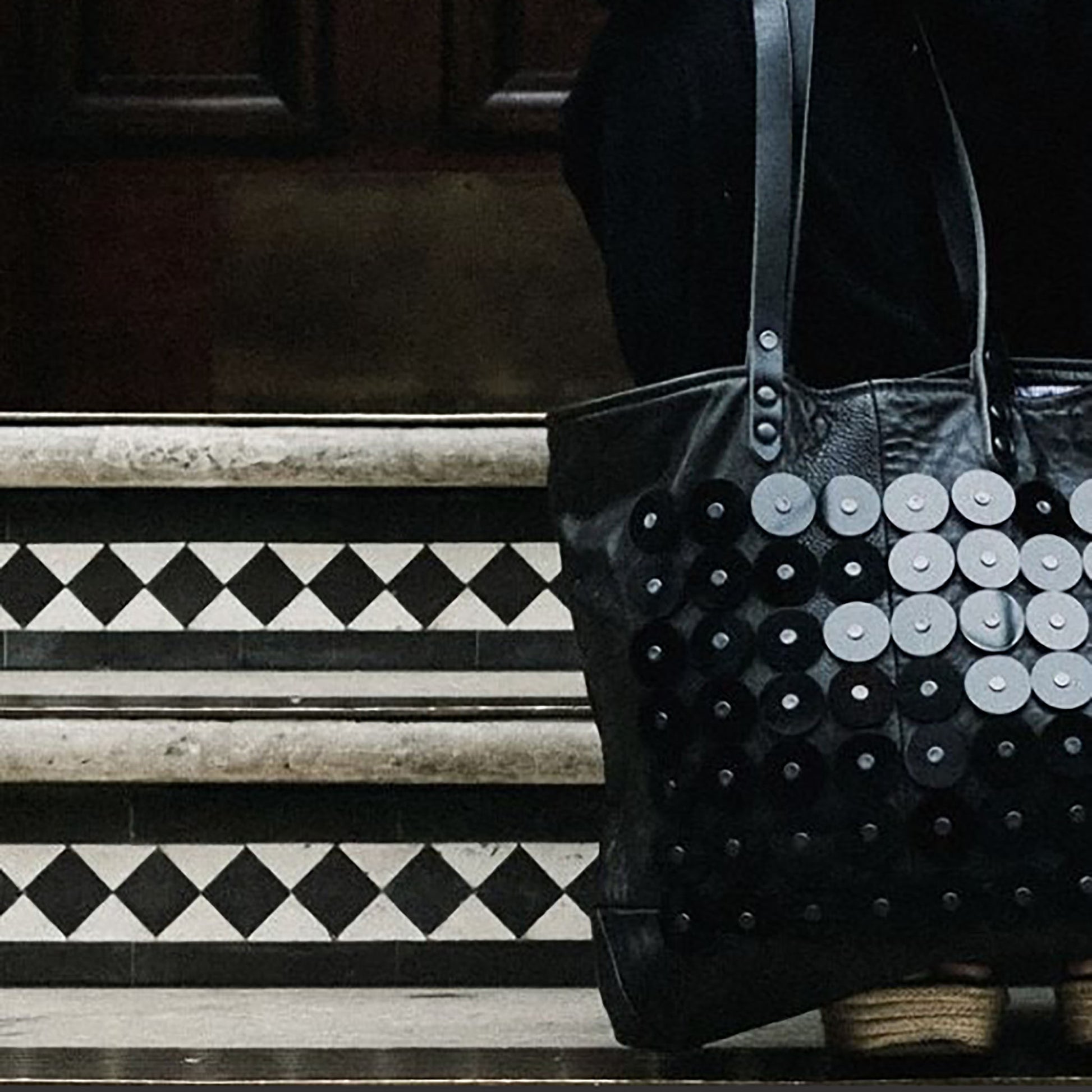 METANoIA black recycled leather medium handbag with black circle acrylic forms fashioned into a repeative fashion with a smaller circle overlay on each form.  Close up on bag infront of geometric tiled stairs.