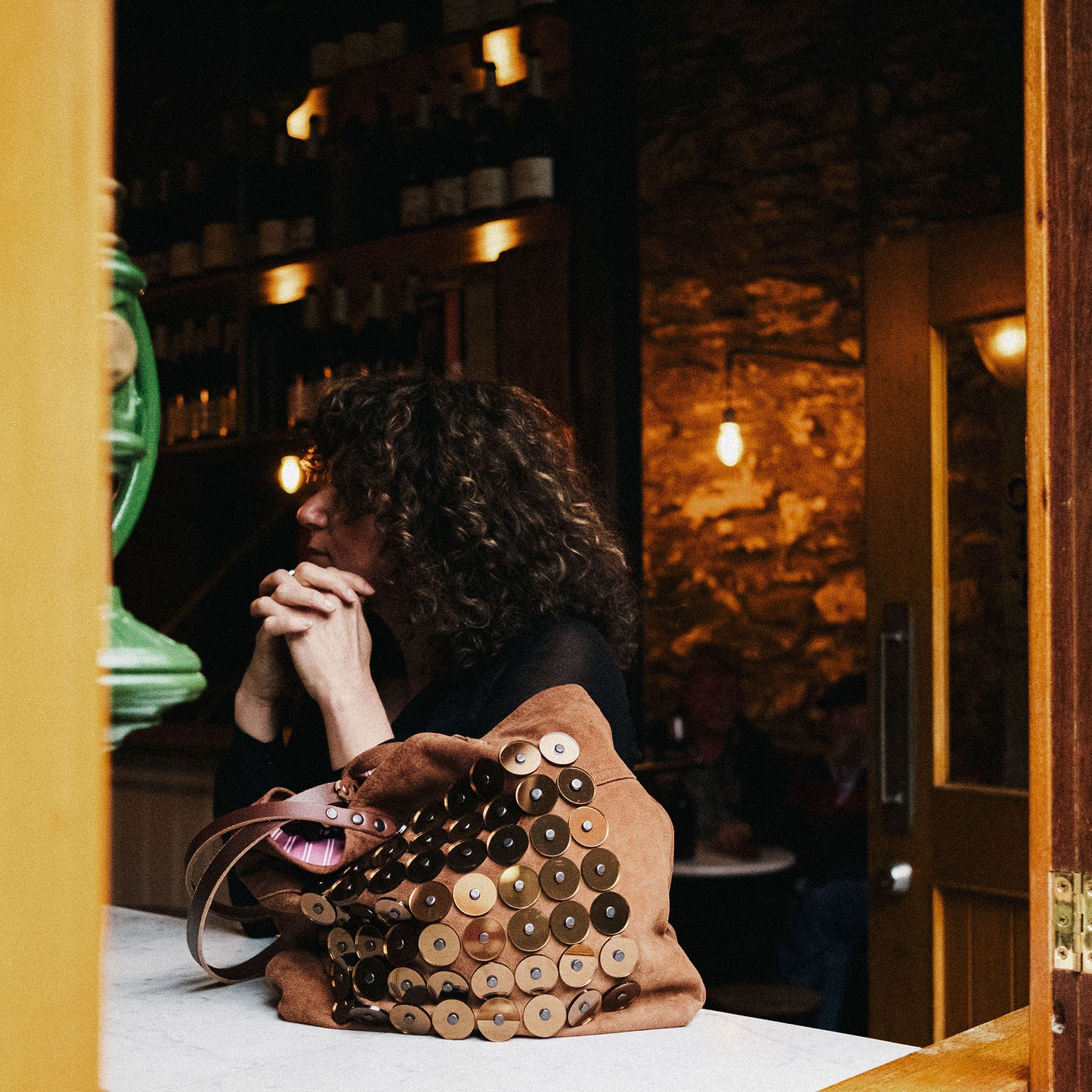 METANoIA tan recycled leather medium handbag with circle bamboo, walnut and acrylic forms fashioned into a repetitive fashion with a smaller circle overlay on each form. Model featured looking into distance with the disco copper bag placed on the marble bar benchtop.