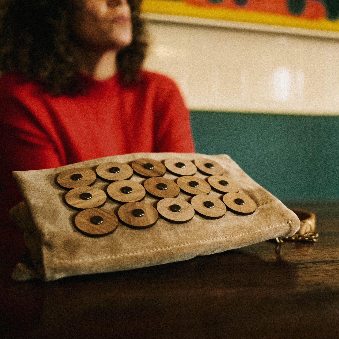 METANoIA tan recycled leather small handbag with circle bamboo and walnut acrylic forms fashioned into a repeative fashion with a smaller circle overlay on each form. Model blurred in background with the small disco bag placed in focus in front, laid down on a table.