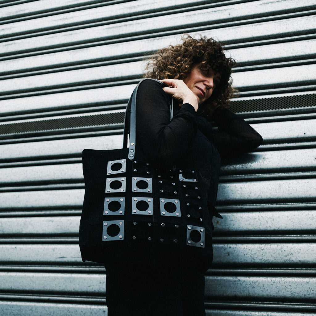METANoIA black recycled leather medium handbag with square and hollowed circle black acrylic forms fashioned into a repetitive fashion. Model featured leaning on industrial metal wall with bag over her shoulder.