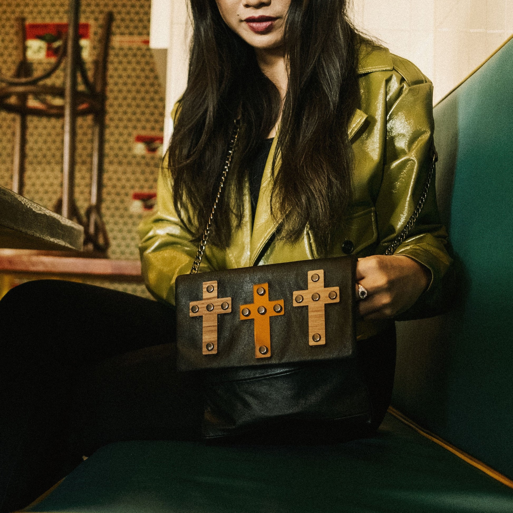 METANoIA The Cross Chocolate recycled leather small handbag with cross shaped bamboo, walnut and metallic acrylic forms. Model featured with cross bag on green vinyl seating.