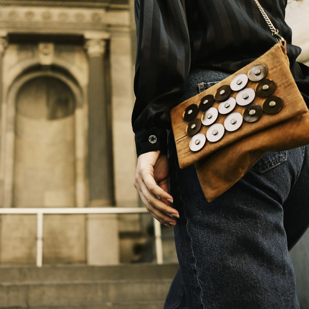 METANoIA tan recycled leather small handbag with circle copper acrylic forms fashioned into a repeative fashion with a smaller circle overlay on each form.  Model featured with bag slung over her shoulder facing a memorial sculpture.