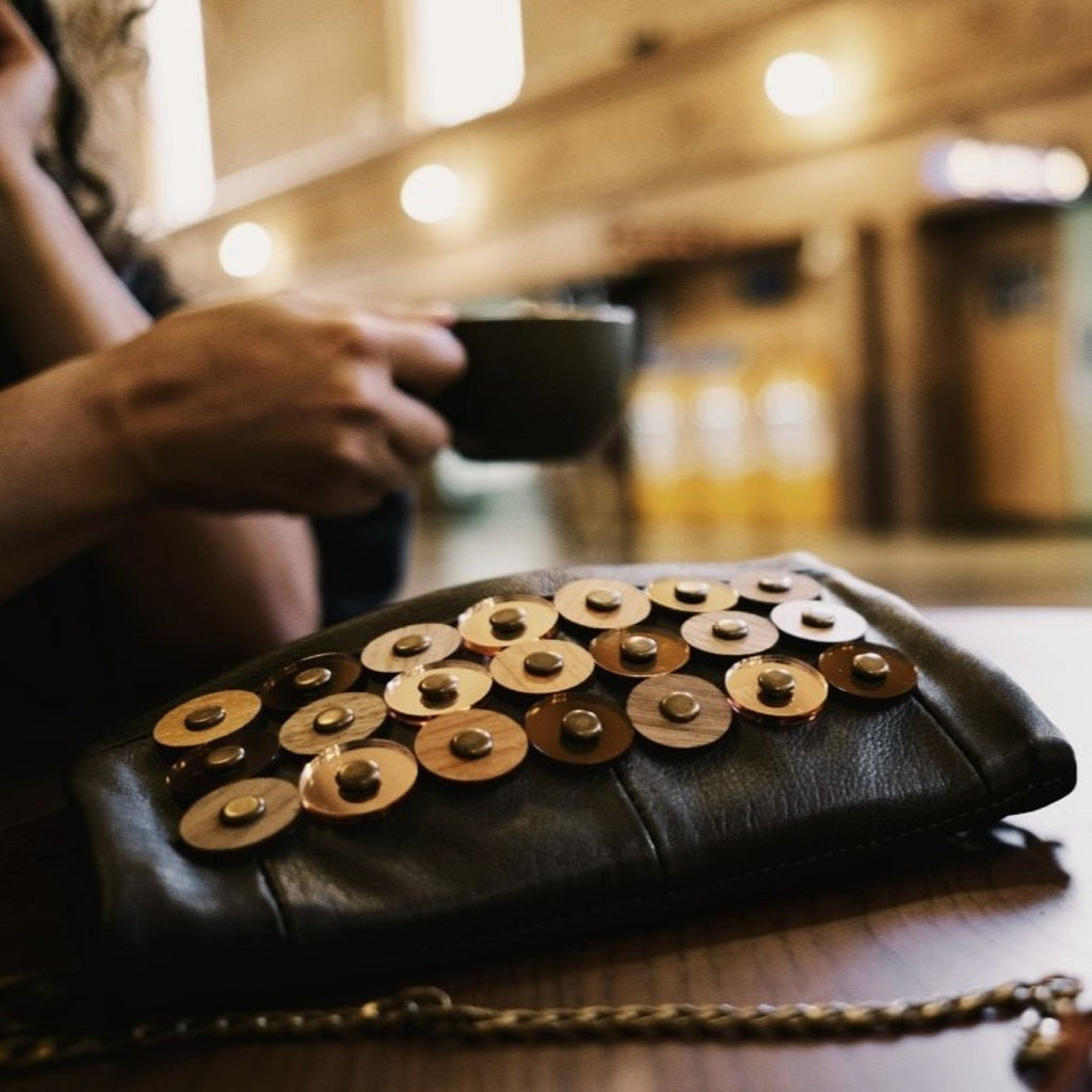 METANoIA black recycled leather small handbag with  circle bamboo, walnut and acrylic forms fashioned into a repeative fashion with a smaller circle overlay on each form. Model featured holding a cup of coffee with the small iris bag beside her.