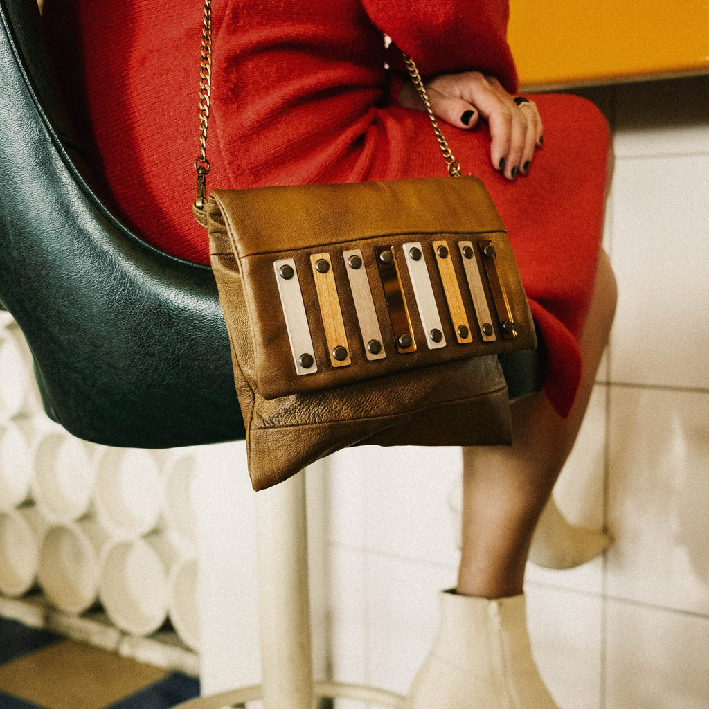 METANoIA tan recycled leather small handbag with vertical rectangular bamboo, walnut, metallic acrylic forms fashioned into a repeative fashion. Model featured on vintage swivel chair wearing a red wool knit dress and white boots with bag slung over her shoulder.