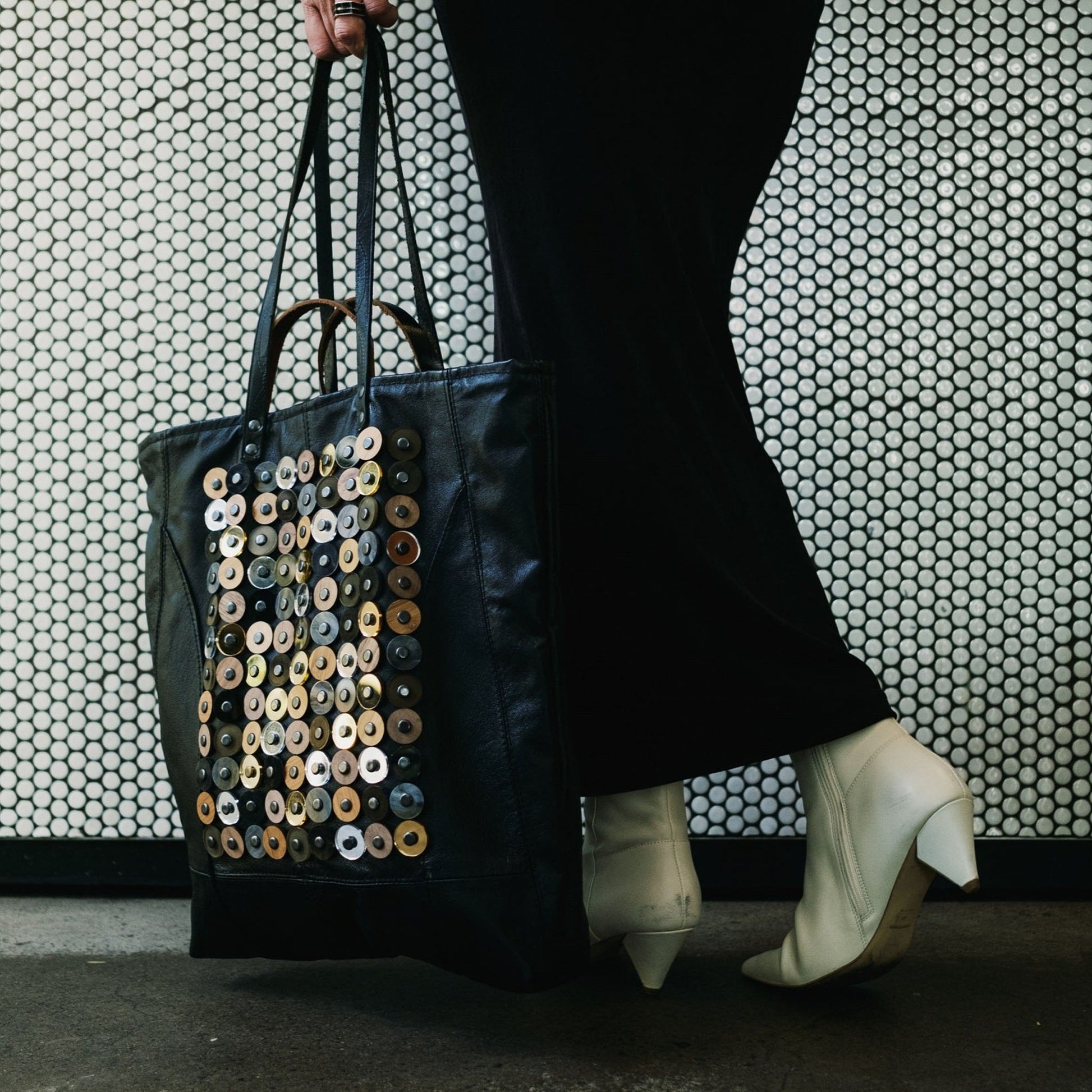 METANoIA black recycled leather tote handbag with  circle bamboo, walnut and acrylic forms fashioned into a repeative fashion with a smaller circle overlay on each form.  Model holding the bag by her side infront of a white mosaic bar.