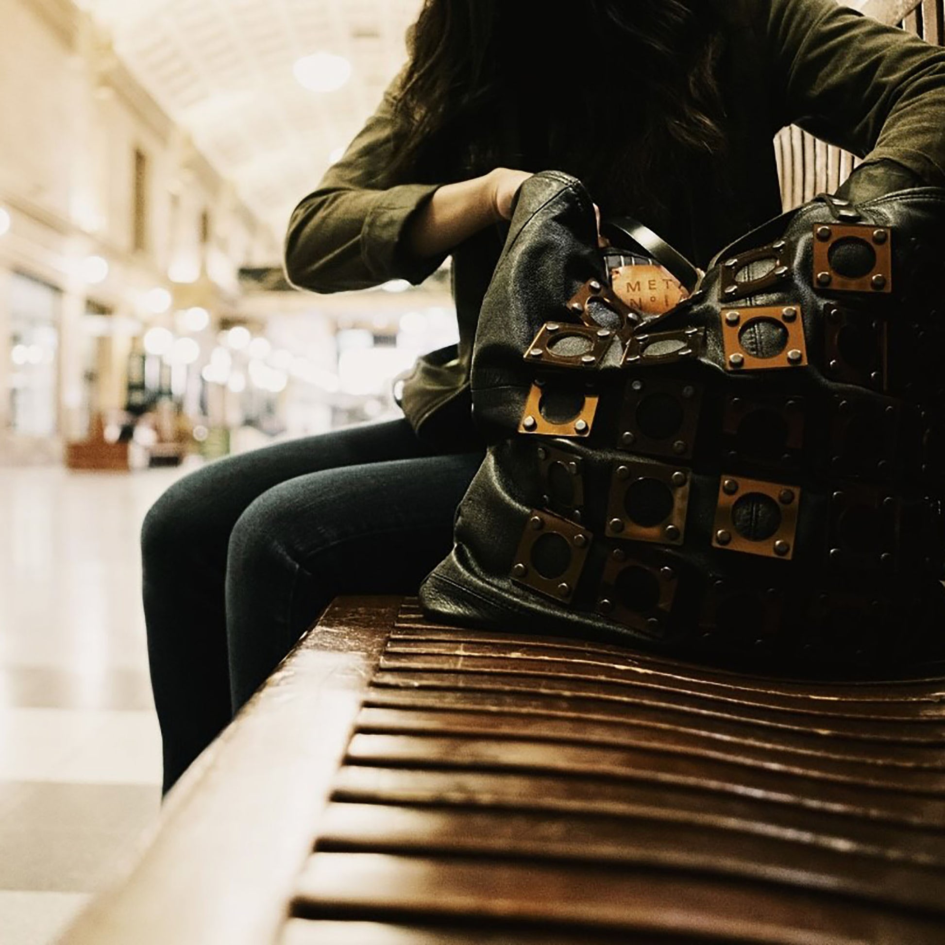 METANoIA black recycled leather tote handbag with square and hollowed circle copper acrylic forms fashioned into a repeative fashion. Model seated on train station bench reaching into the tote bag.