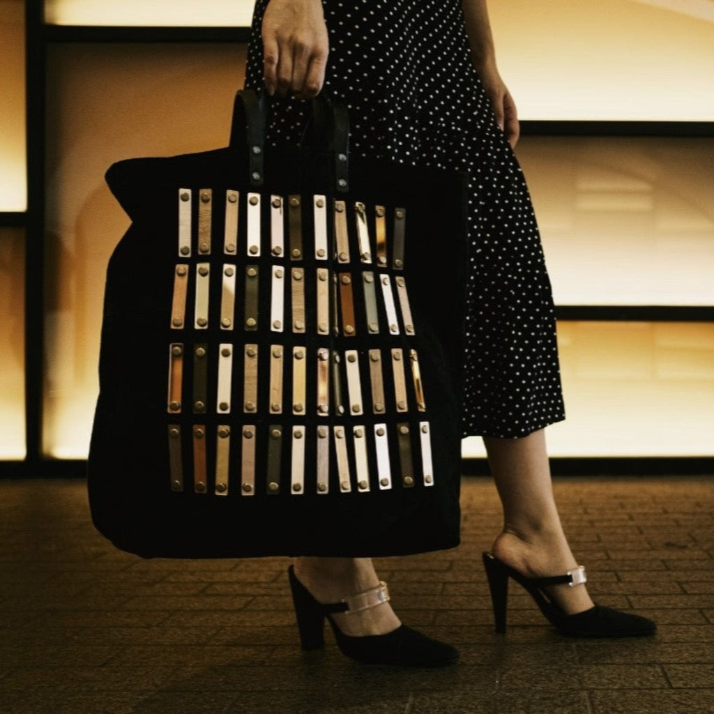METANoIA black recycled leather tote handbag with acrylic and wooden vertical accents studded in a repeatitive fashion in an array of natural wooden browns, coppers and white. Model pictured holding the strokes tote by her side. 