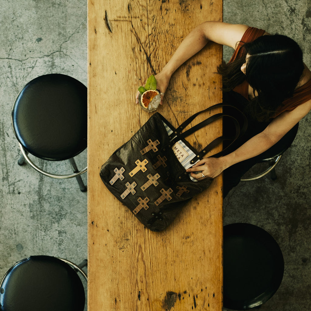 METANoIA The Cross Chocolate recycled leather medium handbag with cross shaped bamboo, walnut and metallic acrylic forms. Model featured with gin in hand and bag placed on a long vintage wooden bench. Photo taken in birdseye view. 