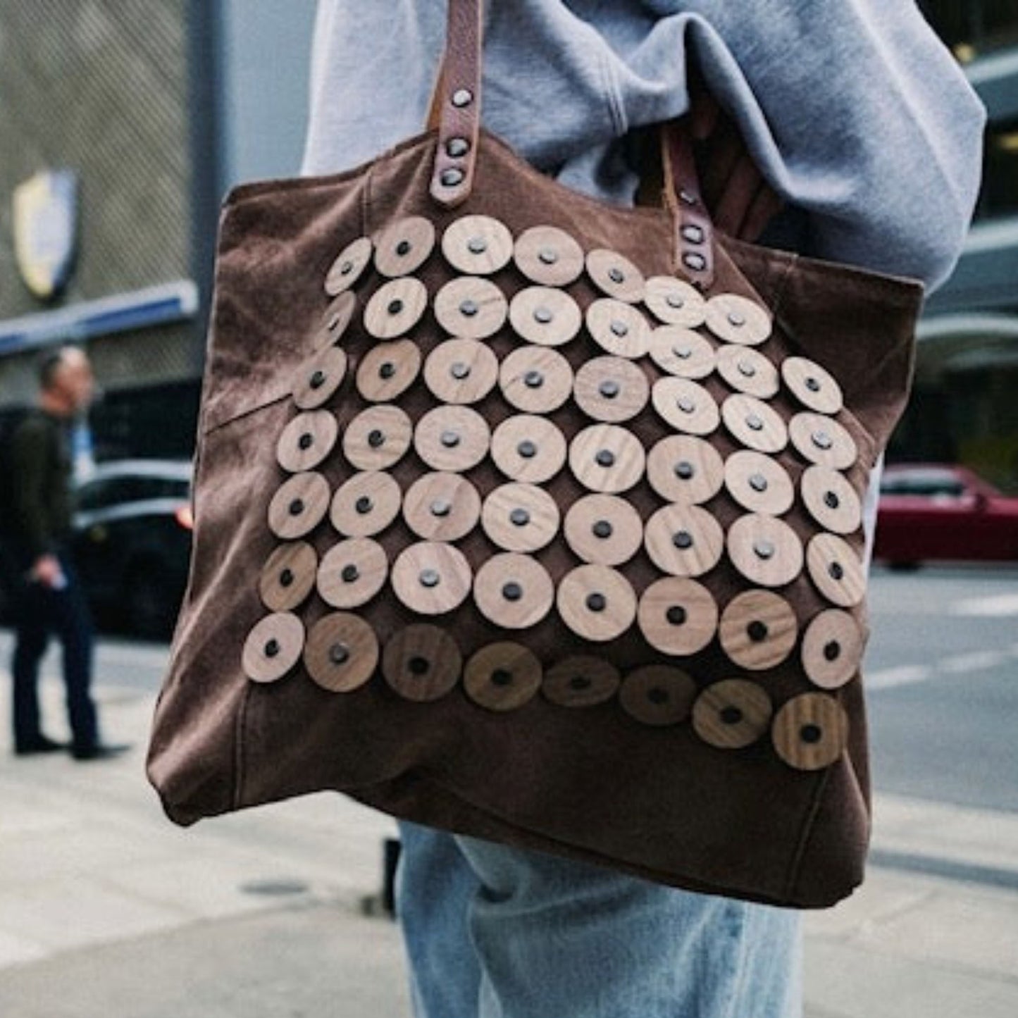 METANoIA light tan recycled leather medium handbag with circle bamboo and walnut forms fashioned into a repetitive fashion with a smaller circle overlay on each form. Model wearing bag over her shoulder while waiting for pedestrian lights.