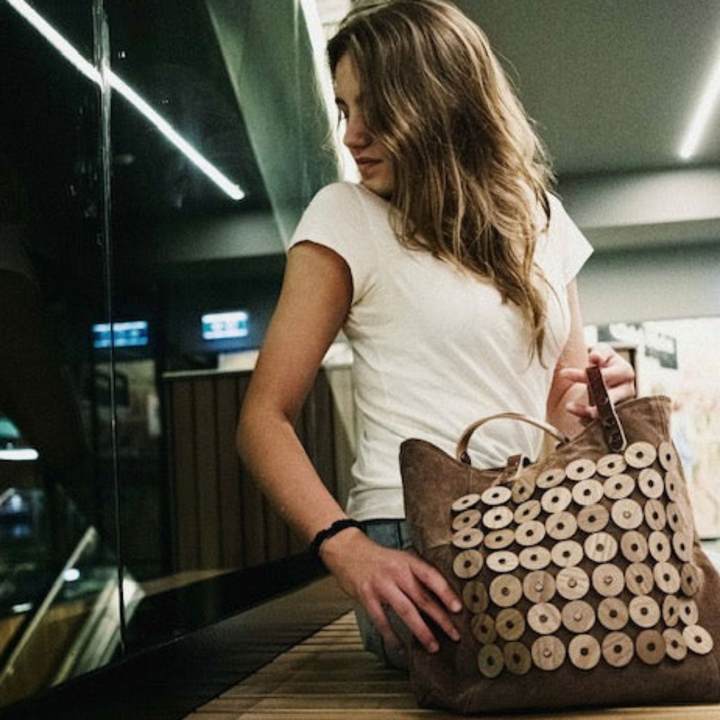 METANoIA light tan recycled leather medium handbag with circle bamboo and walnut forms fashioned into a repeative fashion with a smaller circle overlay on each form. Model staring into distance with bag placed on a wooden bench,