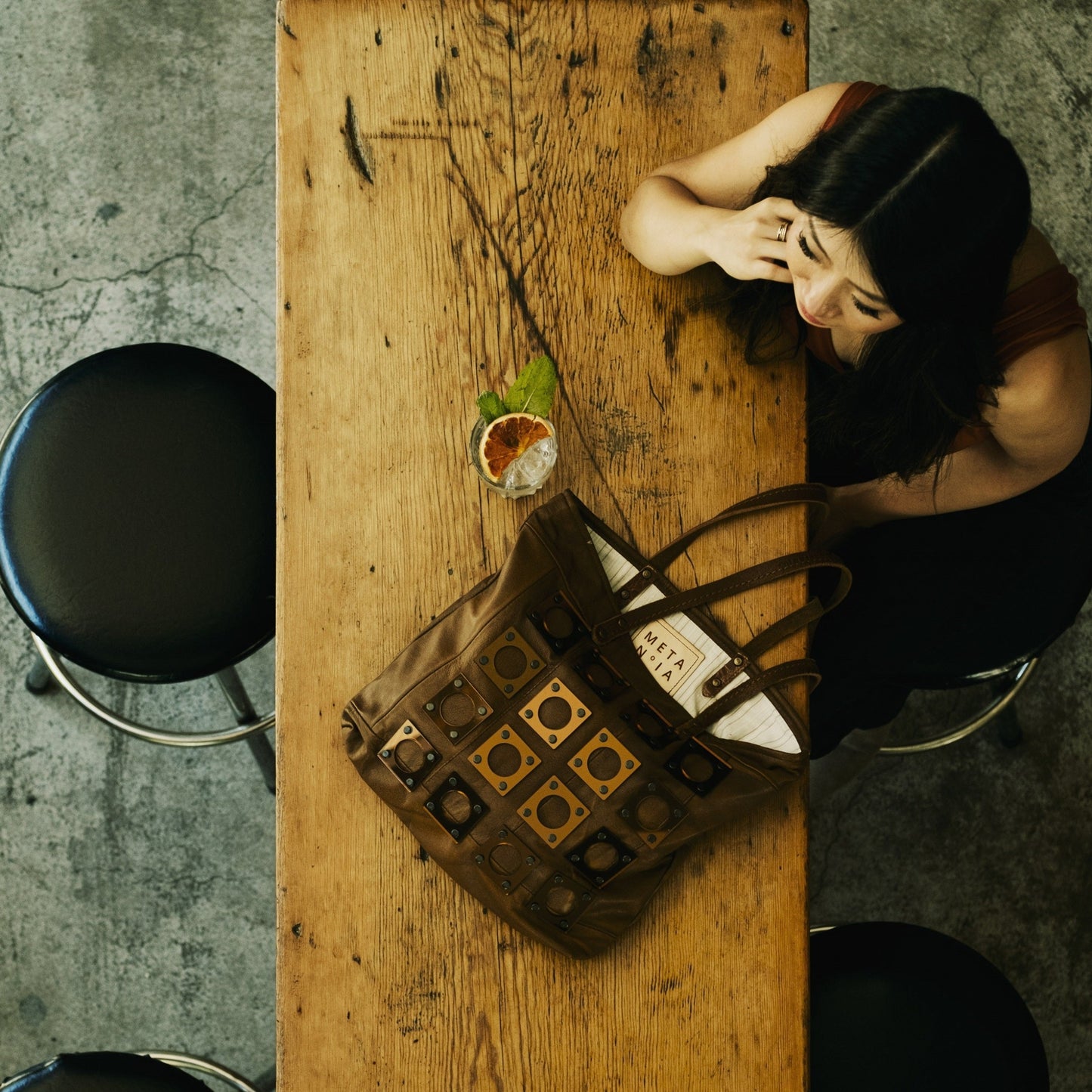 METANoIA caramel recycled leather medium handbag with square and hollowed circle copper acrylic forms fashioned into a repeative fashion. Model featured leaning over wooden bench with gin by her side and bag taken in birdseye view.